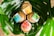 Three wooden soap dishes are nestled within large green leaves. Each dish holds a brightly colored soap package with labels in shades of blue, yellow, red, and green.