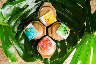 Three wooden soap dishes are nestled within large green leaves. Each dish holds a brightly colored soap package with labels in shades of blue, yellow, red, and green.
