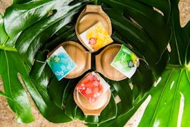 Three wooden soap dishes are nestled within large green leaves. Each dish holds a brightly colored soap package with labels in shades of blue, yellow, red, and green.