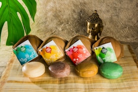 Three bars of soap are displayed on a natural fiber cloth next to a small Buddha statue. Each soap bar is in front of a wooden holder with a colorful and distinct tag: blue for Yogi Soap, orange for Curcuma Soap, and green for Rainforest Soap. A large green leaf is visible on the left side, adding a touch of nature to the setting.