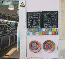 A shopfront displays two chalkboards advertising original tiles and handmade ceramics. The boards are adorned with colorful, intricate tile designs. Below them, two ornate ceramic plates with vibrant, circular patterns are mounted on a pale wall. Inside the shop, shelves are filled with a variety of ceramics and decorative plates.