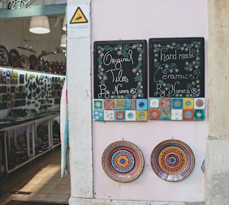 A shopfront displays two chalkboards advertising original tiles and handmade ceramics. The boards are adorned with colorful, intricate tile designs. Below them, two ornate ceramic plates with vibrant, circular patterns are mounted on a pale wall. Inside the shop, shelves are filled with a variety of ceramics and decorative plates.