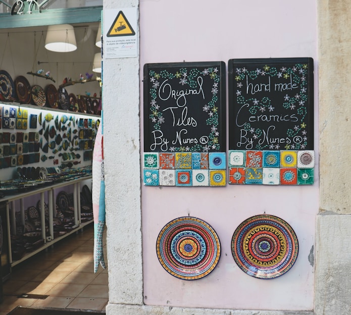 A shopfront displays two chalkboards advertising original tiles and handmade ceramics. The boards are adorned with colorful, intricate tile designs. Below them, two ornate ceramic plates with vibrant, circular patterns are mounted on a pale wall. Inside the shop, shelves are filled with a variety of ceramics and decorative plates.