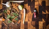 A wooden cutting board hosts a variety of spices and herbs including garlic, lime, cinnamon sticks, nutmeg, cloves, cardamom pods, mint leaves, and turmeric. To the right, there is a bottle labeled 'spice drops' with a dropper cap.