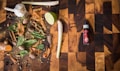 A wooden cutting board hosts a variety of spices and herbs including garlic, lime, cinnamon sticks, nutmeg, cloves, cardamom pods, mint leaves, and turmeric. To the right, there is a bottle labeled 'spice drops' with a dropper cap.