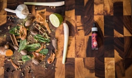 A wooden cutting board hosts a variety of spices and herbs including garlic, lime, cinnamon sticks, nutmeg, cloves, cardamom pods, mint leaves, and turmeric. To the right, there is a bottle labeled 'spice drops' with a dropper cap.