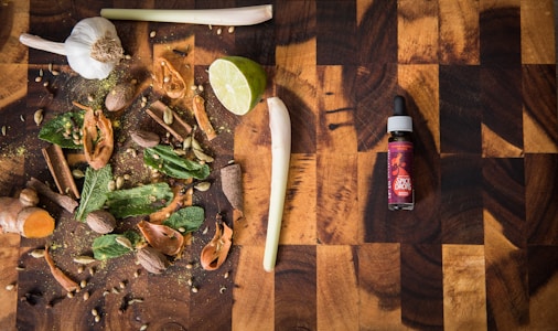 A wooden cutting board hosts a variety of spices and herbs including garlic, lime, cinnamon sticks, nutmeg, cloves, cardamom pods, mint leaves, and turmeric. To the right, there is a bottle labeled 'spice drops' with a dropper cap.
