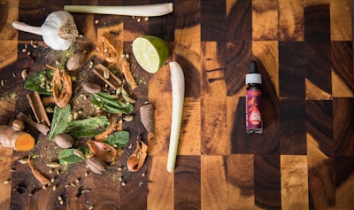 A wooden cutting board hosts a variety of spices and herbs including garlic, lime, cinnamon sticks, nutmeg, cloves, cardamom pods, mint leaves, and turmeric. To the right, there is a bottle labeled 'spice drops' with a dropper cap.