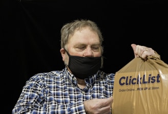 A person wearing a black face mask holds a brown plastic bag with the words 'ClickList Order Online... PICK UP at Store.' The individual is dressed in a blue and white plaid shirt and has short, slightly graying hair. The background is dark, providing contrast to the subject.