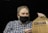 A person wearing a black face mask holds a brown plastic bag with the words 'ClickList Order Online... PICK UP at Store.' The individual is dressed in a blue and white plaid shirt and has short, slightly graying hair. The background is dark, providing contrast to the subject.
