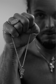 A man with a focused expression holds up a chain with a pendant. His fist prominently displays a ring with an emblem. A tattoo on his forearm reads 'CREATOR.' The monochromatic setting adds intensity to the scene.