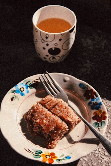 A cup filled with a warm-orange colored beverage sits on a dark surface, next to a floral decorated plate holding three pieces of a crumbly dessert with a fork resting beside them.