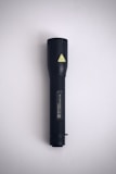 A black flashlight is positioned vertically against a plain, light-gray background. It features a textured grip and a small yellow caution symbol near the top.