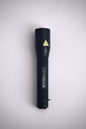 A black flashlight is positioned vertically against a plain, light-gray background. It features a textured grip and a small yellow caution symbol near the top.