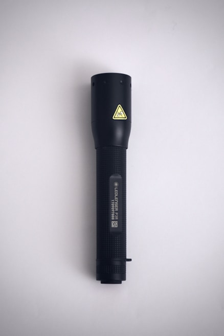 A black flashlight is positioned vertically against a plain, light-gray background. It features a textured grip and a small yellow caution symbol near the top.