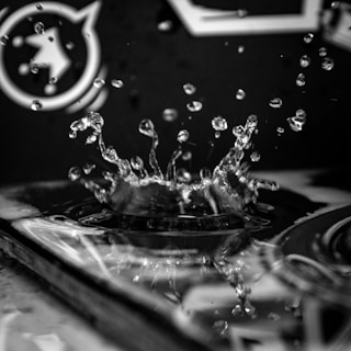 A high-contrast black and white image capturing a water droplet splash in sharp detail. The droplets appear suspended in the air around the point of impact, creating a dynamic and energetic scene. The background is slightly blurred, focusing attention on the water's motion.