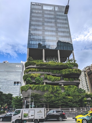 A modern urban building features lush, green terraces on multiple levels, integrating nature into an urban environment. The upper floors consist of a sleek, glass structure, creating a striking contrast with the greenery below. Several vehicles, including cars and a delivery truck, are visible in the foreground, indicating a busy city street. Nearby, another tall building is visible, characterized by a simple, geometric design.