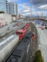 A red and black freight train travels on railway tracks running parallel to a busy urban street. High-rise buildings and construction cranes are visible, along with a bridge over a river in the background. Several cars and trucks are parked or moving along the adjacent street.