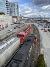 A red and black freight train travels on railway tracks running parallel to a busy urban street. High-rise buildings and construction cranes are visible, along with a bridge over a river in the background. Several cars and trucks are parked or moving along the adjacent street.