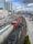 A red and black freight train travels on railway tracks running parallel to a busy urban street. High-rise buildings and construction cranes are visible, along with a bridge over a river in the background. Several cars and trucks are parked or moving along the adjacent street.