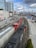 A red and black freight train travels on railway tracks running parallel to a busy urban street. High-rise buildings and construction cranes are visible, along with a bridge over a river in the background. Several cars and trucks are parked or moving along the adjacent street.