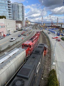 A red and black freight train travels on railway tracks running parallel to a busy urban street. High-rise buildings and construction cranes are visible, along with a bridge over a river in the background. Several cars and trucks are parked or moving along the adjacent street.