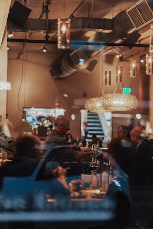 A cozy and dimly lit bar or restaurant with a warm ambiance where people are gathered around a bar counter. Pendant lights hang from the ceiling, and there is visible industrial-style ductwork. The image shows various bottles and glasses, and a bartender serving drinks as patrons sit and converse.