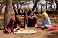 Three individuals are seated on the ground in a forested area with sparse trees and dry grass. Two of them are wearing traditional Maasai attire, characterized by red and blue plaid shawls. A Caucasian woman in a light blue shirt and beige pants sits on a red blanket. One of the Maasai individuals appears to be engaging in a traditional activity involving a large gourd, which could be related to food preparation or ritual. In the background, there are more people dressed similarly and a herd of cattle.