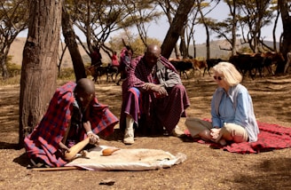 Three individuals are seated on the ground in a forested area with sparse trees and dry grass. Two of them are wearing traditional Maasai attire, characterized by red and blue plaid shawls. A Caucasian woman in a light blue shirt and beige pants sits on a red blanket. One of the Maasai individuals appears to be engaging in a traditional activity involving a large gourd, which could be related to food preparation or ritual. In the background, there are more people dressed similarly and a herd of cattle.