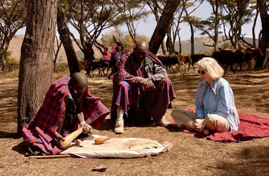 Three individuals are seated on the ground in a forested area with sparse trees and dry grass. Two of them are wearing traditional Maasai attire, characterized by red and blue plaid shawls. A Caucasian woman in a light blue shirt and beige pants sits on a red blanket. One of the Maasai individuals appears to be engaging in a traditional activity involving a large gourd, which could be related to food preparation or ritual. In the background, there are more people dressed similarly and a herd of cattle.