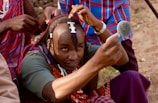 A person carefully styling their hair using a small mirror, with assistance from another individual behind them. The person holds a mirror close to their face while adjusting dreadlocks adorned with white beads, against the backdrop of traditional clothing.