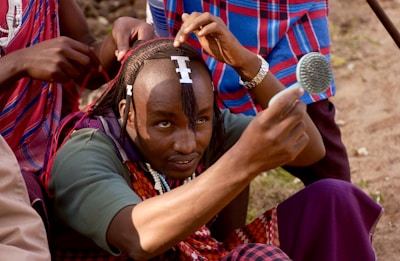 A person carefully styling their hair using a small mirror, with assistance from another individual behind them. The person holds a mirror close to their face while adjusting dreadlocks adorned with white beads, against the backdrop of traditional clothing.