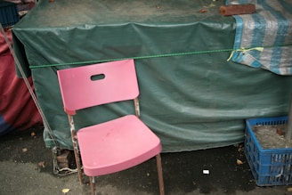 A pink plastic chair is placed in front of a green tarpaulin-covered structure. The chair is slightly worn and stands on an asphalt surface. To the right, there is a blue plastic crate, and a striped blue and white cloth is draped over the green tarp. A rope is tied around the structure, adding to the makeshift appearance.