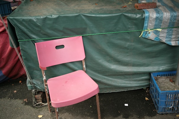 A pink plastic chair is placed in front of a green tarpaulin-covered structure. The chair is slightly worn and stands on an asphalt surface. To the right, there is a blue plastic crate, and a striped blue and white cloth is draped over the green tarp. A rope is tied around the structure, adding to the makeshift appearance.