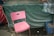 A pink plastic chair is placed in front of a green tarpaulin-covered structure. The chair is slightly worn and stands on an asphalt surface. To the right, there is a blue plastic crate, and a striped blue and white cloth is draped over the green tarp. A rope is tied around the structure, adding to the makeshift appearance.
