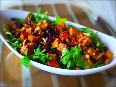 A vibrant salad is presented in an elongated white dish, featuring fresh green leaves, crispy croutons, dried cranberries, and sliced almonds. The ingredients are artistically arranged, showcasing a mix of textures and colors.