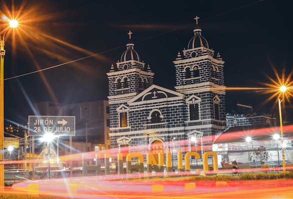 A nighttime scene depicting a well-lit church with twin towers adorned with crosses. The building features a striking black and white facade that is illuminated against the dark sky. In the foreground, there is a sign pointing towards 'Jirón 28 de Julio,' and large letters spelling out 'Huánuco.' Long exposure light trails from passing vehicles add dynamic movement to the scene.