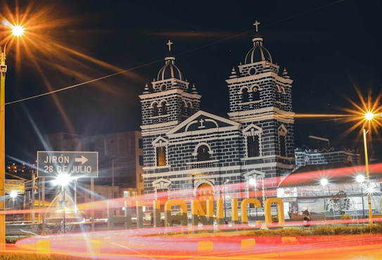 A nighttime scene depicting a well-lit church with twin towers adorned with crosses. The building features a striking black and white facade that is illuminated against the dark sky. In the foreground, there is a sign pointing towards 'Jirón 28 de Julio,' and large letters spelling out 'Huánuco.' Long exposure light trails from passing vehicles add dynamic movement to the scene.
