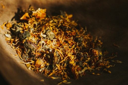 A collection of dried flowers and leaves is gathered in a wooden bowl. The arrangement includes orange and brown tones, hinting at a natural and rustic setting. The lighting creates a warm and soft atmosphere, with shadows adding depth and contrast.