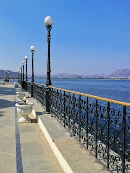 A scenic view of a lakeside promenade with ornate black iron railings and vintage-style street lamps. The promenade features large, decorative stone planters. In the background, a calm lake stretches towards distant hills under a clear blue sky.