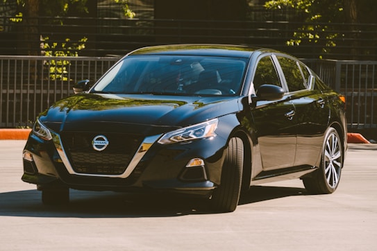 A sleek, black sedan is parked in an outdoor setting, with sunlight reflecting off its shiny surface. The car features modern design elements, such as sharp headlights, a prominent grille, and alloy wheels.
