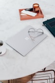 A minimalistic workspace setup featuring a closed laptop with a pair of glasses resting on top, a burning candle in a brown glass jar, a tray holding a few notebooks, and a cup with a beverage. These items are arranged on a round, white marble table with a simple, clean aesthetic.