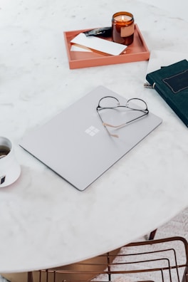 A minimalistic workspace setup featuring a closed laptop with a pair of glasses resting on top, a burning candle in a brown glass jar, a tray holding a few notebooks, and a cup with a beverage. These items are arranged on a round, white marble table with a simple, clean aesthetic.