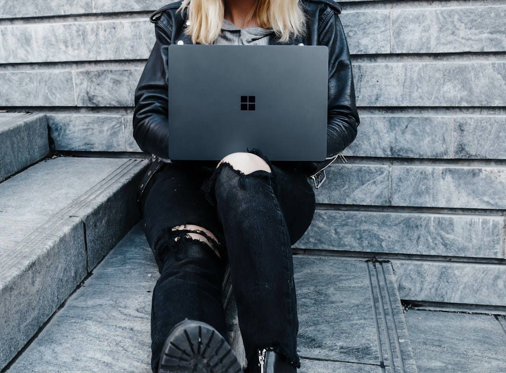 woman in black jacket and black pants sitting on concrete stairs usingMicrosoft Surface Laptop  
