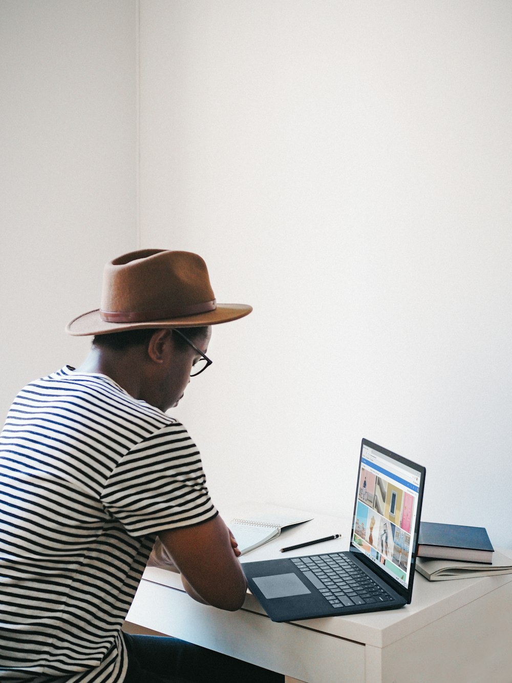 man in white and black stripe shirt wearing brown hat sitting on chair using a Microsoft surface cobalt laptop 