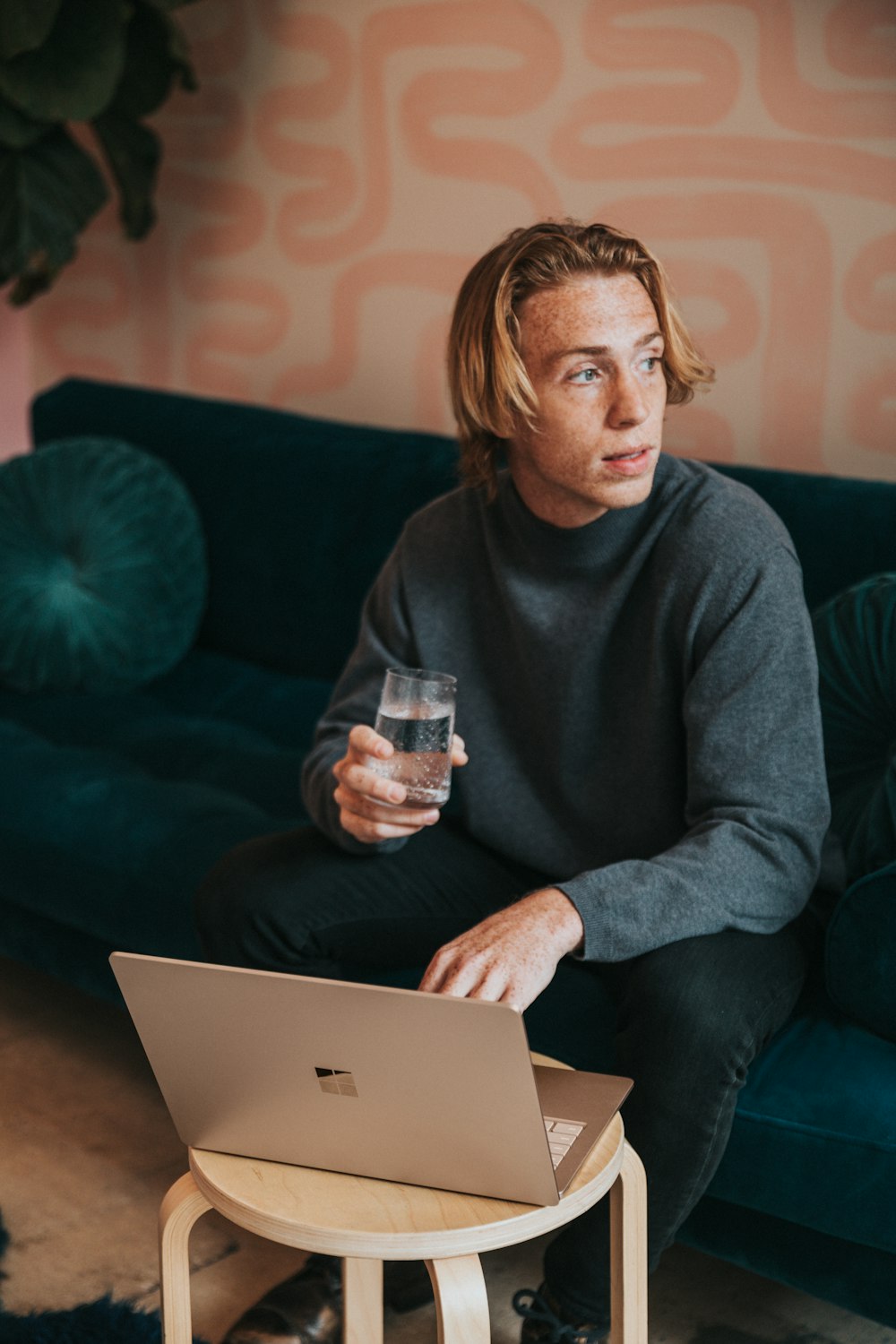 man in gray sweater sitting on blue couch holding clear drinking glass working on a Surface 3 Sandstone laptop 