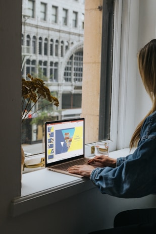 A person in a denim jacket is sitting by a windowsill using a laptop. The laptop screen displays a presentation titled 'Travel Inspiration' with a colorful template featuring triangles and a building. Outside the window, an urban street scene with an arched building facade is visible.