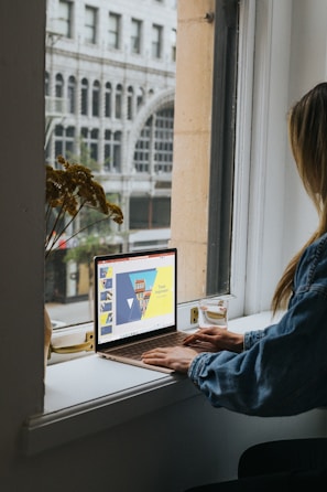 A person in a denim jacket is sitting by a windowsill using a laptop. The laptop screen displays a presentation titled 'Travel Inspiration' with a colorful template featuring triangles and a building. Outside the window, an urban street scene with an arched building facade is visible.