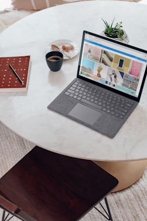 A modern workspace is arranged on a round marble table, featuring a sleek laptop displaying various images on its screen. Nearby, a black cup filled with coffee sits beside a small plate of biscuits. A closed notebook with a geometric pattern and a pen lay on the table. An open plant pot adds a touch of greenery to the setup. A wooden chair is positioned next to the table over a textured carpet.