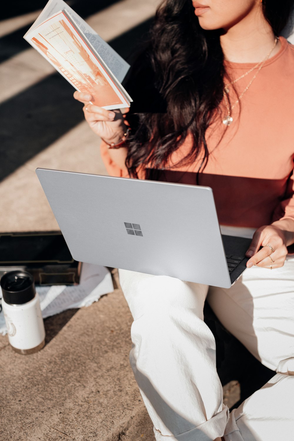 オレンジ色のシャツと白いズボンを着た女性が銀色のMicrosoft Surfaceラップトップコンピューターを持っている